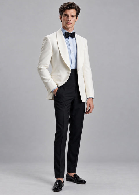A man modeling a white tuxedo jacket with black shawl lapels, paired with black trousers, a light blue dress shirt, a black bow tie, and black loafers, against a solid gray studio background.