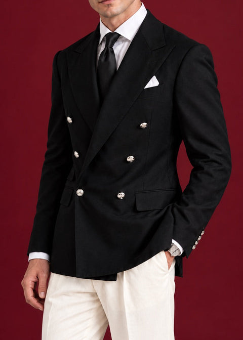 A man modeling a black double-breasted blazer with silver-tone buttons, paired with a white dress shirt, black tie, and white trousers, set against a solid dark red studio background.