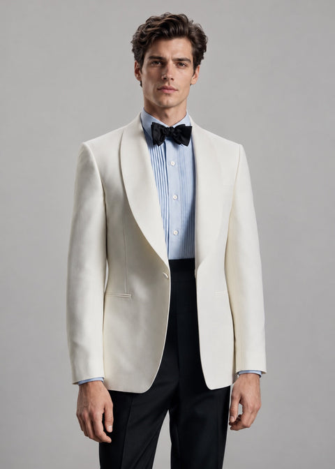 A man modeling a white tuxedo jacket with peak lapels, worn over a light blue striped dress shirt, a black bow tie, and black trousers, against a solid gray studio background.