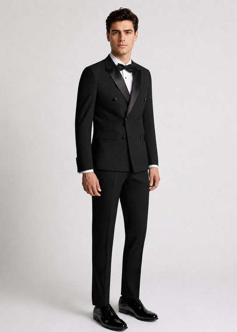 A man modeling a classic black double-breasted tuxedo with satin lapels, a white dress shirt, a black bow tie, and black dress shoes against a light gray studio background.
